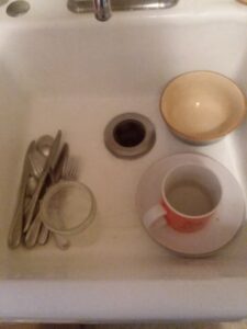 A sink with lots of empty space, a mug of silverware, one bowl, two plates, one glass jar and a cup. 