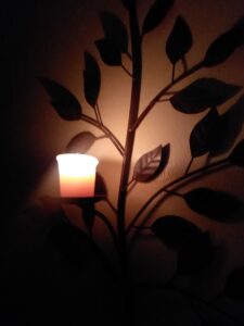 A small candle burns on a metal candle holder on a wall. The candleholder is shaped like a branch with leaves. 